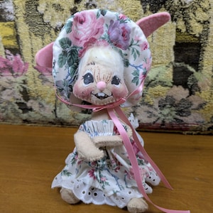Puede incluir: Mu&ntilde;eca de conejito hecha a mano con un gorro y un vestido florales. El gorro presenta rosas rosas y moradas sobre un fondo blanco. El vestido tiene una falda calada blanca y una cinta rosa. La mu&ntilde;eca tiene una cara sonriente y orejas rosas.