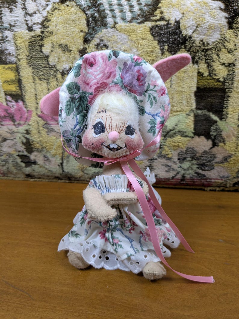 Puede incluir: Mu&ntilde;eca de conejito hecha a mano con un gorro y un vestido florales. El gorro presenta rosas rosas y moradas sobre un fondo blanco. El vestido tiene una falda calada blanca y una cinta rosa. La mu&ntilde;eca tiene una cara sonriente y orejas rosas.