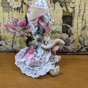 Puede incluir: Una mu&ntilde;eca de conejito de peluche hecha a mano con un vestido y un gorro con estampado floral. El vestido presenta rosas rosas y un dobladillo de encaje calado blanco. El conejito tiene orejas color canela y un lazo rosa.