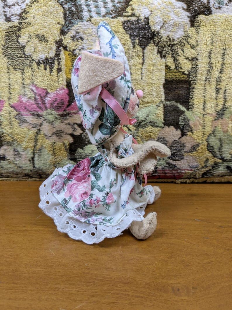 Puede incluir: Una mu&ntilde;eca de conejito de peluche hecha a mano con un vestido y un gorro con estampado floral. El vestido presenta rosas rosas y un dobladillo de encaje calado blanco. El conejito tiene orejas color canela y un lazo rosa.
