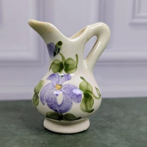 May include: A small cream-colored ceramic pitcher featuring a hand-painted purple flower design with green leaves. The pitcher has a curved handle, a flared spout, and a rounded base, showcasing a speckled finish throughout the ceramic surface.