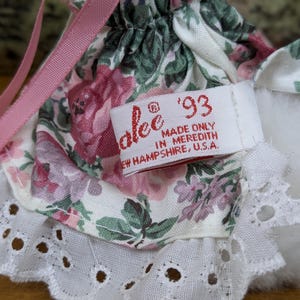 Puede incluir: Primer plano de un vestido de mu&ntilde;eca con un estampado floral en rosa, verde y blanco. El vestido tiene un ribete de encaje blanco y una cinta rosa. Una etiqueta dice "alee '93 MADE ONLY IN MEREDITH NEW HAMPSHIRE, U.S.A."