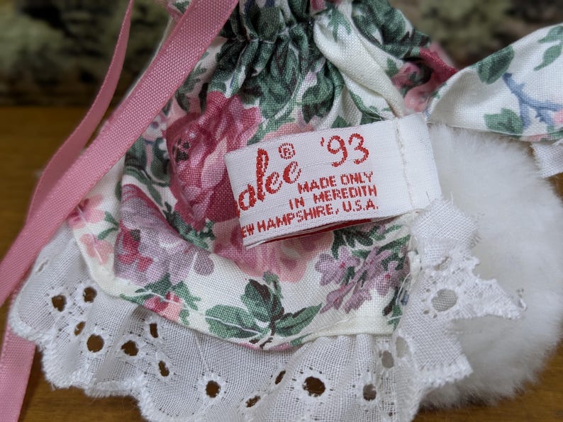 Puede incluir: Primer plano de un vestido de mu&ntilde;eca con un estampado floral en rosa, verde y blanco. El vestido tiene un ribete de encaje blanco y una cinta rosa. Una etiqueta dice "alee '93 MADE ONLY IN MEREDITH NEW HAMPSHIRE, U.S.A."