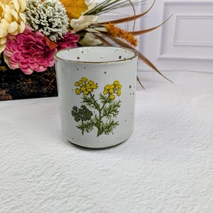 May include: A cylindrical ceramic mug with a speckled off-white glaze and a brown rim. The front features a botanical illustration of yellow flowers with green stems and leaves. The mug has a smooth, straight-sided body and a flat base.