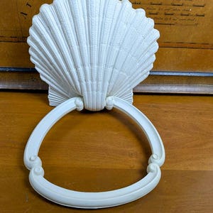May include: A white seashell-shaped wall hanging with a curved, circular handle. The seashell has detailed ridges and is mounted on a rectangular base. The handle has decorative scrollwork on each side.