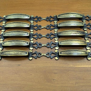 May include: A collection of antique-style cabinet pulls. Each pull features a textured, brass-colored handle with ornate, dark bronze-colored decorative elements. The pulls are arranged in two rows on a wooden surface.