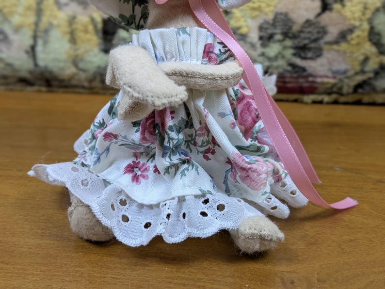 Puede incluir: Una mu&ntilde;eca hecha a mano con un vestido estampado floral con un ribete de encaje de ojal blanco. El vestido presenta rosas rosas y rojas. La mu&ntilde;eca tiene brazos y piernas beige, y una cinta rosa est&aacute; unida al sombrero de la mu&ntilde;eca.
