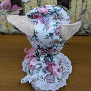 Puede incluir: Un juguete de peluche hecho a mano con una cabeza y orejas con estampado floral, y un vestido blanco con un ribete de encaje. La tela presenta rosas rosas y moradas sobre un fondo blanco. Una cinta rosa est&aacute; atada alrededor del cuello.