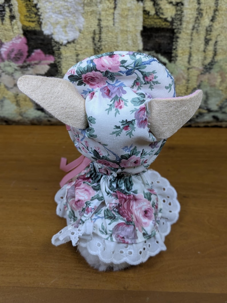 Puede incluir: Un juguete de peluche hecho a mano con una cabeza y orejas con estampado floral, y un vestido blanco con un ribete de encaje. La tela presenta rosas rosas y moradas sobre un fondo blanco. Una cinta rosa est&aacute; atada alrededor del cuello.