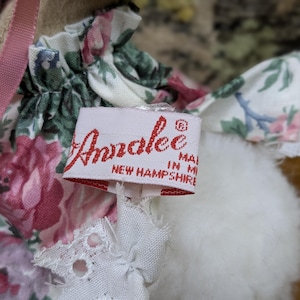 Puede incluir: Primer plano de una mu&ntilde;eca con un vestido floral y una cinta rosa. La mu&ntilde;eca tiene una etiqueta que dice "Annalee Made in New Hampshire" en letras rojas.