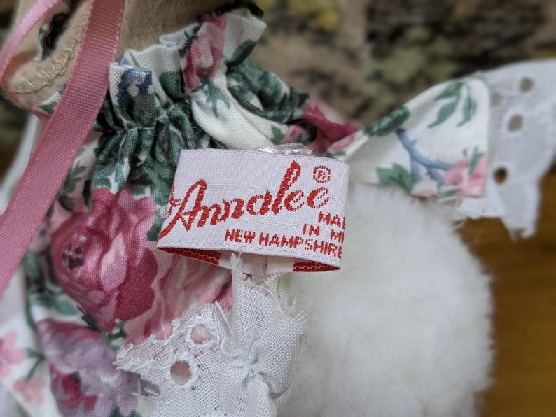 Puede incluir: Primer plano de una mu&ntilde;eca con un vestido floral y una cinta rosa. La mu&ntilde;eca tiene una etiqueta que dice "Annalee Made in New Hampshire" en letras rojas.