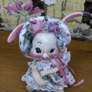 Puede incluir: Una mu&ntilde;eca de conejito hecha a mano con un gorro y un vestido con estampado floral con detalles en rosa. El conejito tiene una cara dulce con ojos negros y una nariz rosa. Las orejas de la mu&ntilde;eca son rosas y tiene un peinado blanco y esponjoso.