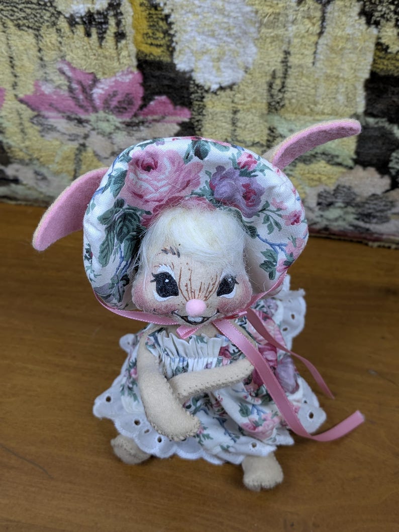Puede incluir: Una mu&ntilde;eca de conejito hecha a mano con un gorro y un vestido con estampado floral con detalles en rosa. El conejito tiene una cara dulce con ojos negros y una nariz rosa. Las orejas de la mu&ntilde;eca son rosas y tiene un peinado blanco y esponjoso.