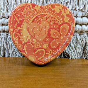 May include: Heart-shaped decorative tin with a vibrant orange and yellow floral design. The tin features a central monogram and the brand name "Brighton". The tin is made in China.