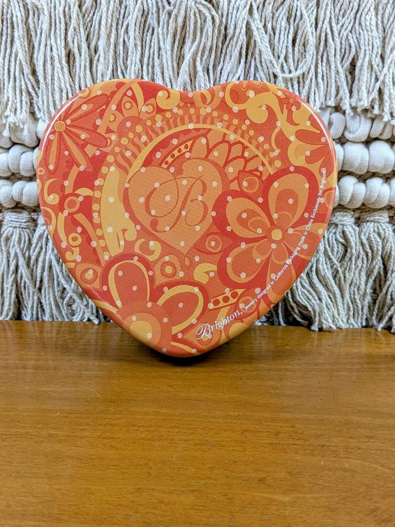 May include: Heart-shaped decorative tin with a vibrant orange and yellow floral design. The tin features a central monogram and the brand name "Brighton". The tin is made in China.