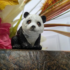 May include: A small panda figurine featuring a white face with black patches around the eyes and ears, a black nose, and brown eyes. The body is primarily black with textured fur detailing, depicted in a seated position with front paws visible.
