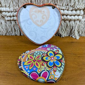 May include: Heart-shaped Brighton trinket box with a floral design lid. The lid features a colorful pattern of flowers in yellow, blue, pink, and orange. The inside of the box is white with a Brighton logo.