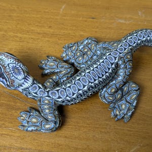May include: A detailed, handcrafted lizard figurine. The sculpture features a mosaic-like pattern in shades of blue, white, and brown. The lizard's body is elongated, with detailed legs and a curved tail, resting on a wooden surface.