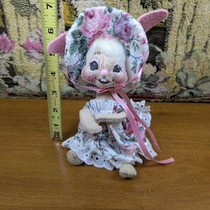 Puede incluir: Una mu&ntilde;eca vintage con cara de conejito, con un gorro y un vestido florales. El gorro y el vestido presentan rosas rosas y moradas sobre un fondo blanco. La mu&ntilde;eca mide aproximadamente 15 cm de altura.