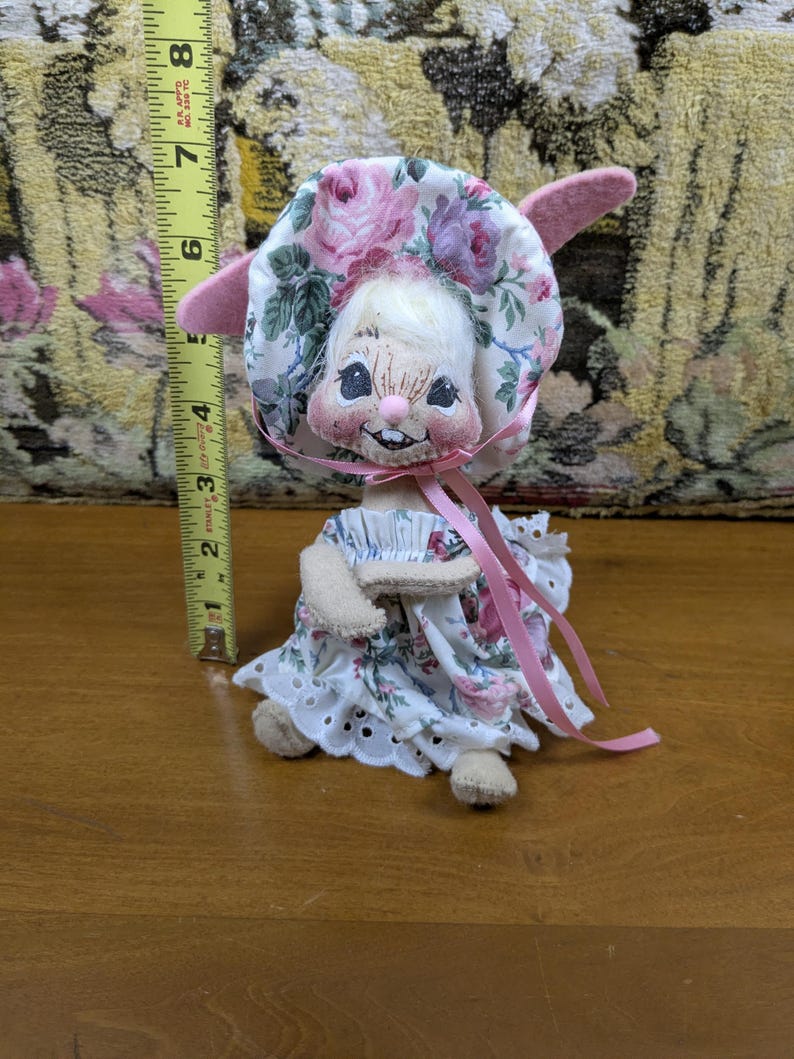 Puede incluir: Una mu&ntilde;eca vintage con cara de conejito, con un gorro y un vestido florales. El gorro y el vestido presentan rosas rosas y moradas sobre un fondo blanco. La mu&ntilde;eca mide aproximadamente 15 cm de altura.