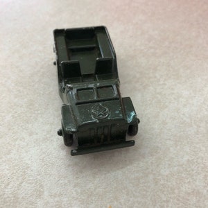 May include: A small olive green die-cast metal toy military vehicle featuring an open-top design, a front bumper, and a molded star emblem on the hood. The vehicle has two front seats and a rear cargo area, with visible paint wear on the edges.