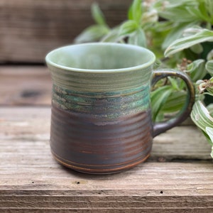 May include: A handcrafted ceramic mug with a green interior and a dark brown exterior. The mug features a textured, ridged design and a curved handle. The mug is placed on a wooden surface.