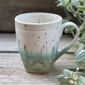 Könnte beinhalten: Eine weiße Keramiktasse mit einem gesprenkelten Überzug und einem grünen und blauen Überzug am Boden. Die Tasse hat einen Henkel und steht auf einer Holzoberfläche.