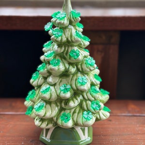 Könnte beinhalten: Ein grüner Keramik-Weihnachtsbaum, verziert mit Kleeblatt-Verzierungen. Der Baum hat eine strukturierte Oberfläche und steht auf einem grünen Sockel mit weißen Akzenten. Der Baum ist etwa 20 cm hoch.