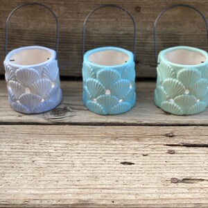 May include: Three ceramic candle holders with a seashell design. The holders are light blue, medium blue, and light green. Each holder has a black metal handle.