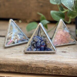 May include: Three glass pyramid-shaped boxes with silver frames. Each box contains different colored stones. The box on the left contains white and brown stones. The box in the middle contains blue stones. The box on the right contains pink stones.