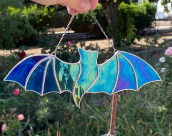 Puede incluir: Un murciélago de vitral colgado de una cadena plateada. El murciélago está hecho de vidrio azul, verde y morado y tiene un contorno plateado. El murciélago está colgado frente a un arbusto verde.