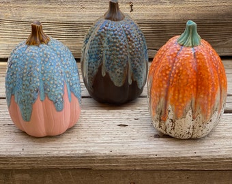 Può includere: Tre zucche in ceramica di diversi colori. Le zucche sono decorate con una glassa macchiettata e hanno un effetto di glassa a goccia. Le zucche sono disposte in fila su una superficie di legno.