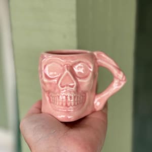 Può includere: Una tazza di ceramica rosa a forma di teschio umano. La tazza ha un manico che fa parte del braccio del teschio. I lineamenti del teschio, comprese le orbite, il naso e i denti, sono ben definiti. La tazza è tenuta in una mano.