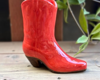 May include: A glossy, red ceramic cowboy boot with a brown heel and sole. The boot is a decorative item, likely a vase or small planter, and is set against a wooden surface with green foliage in the background.
