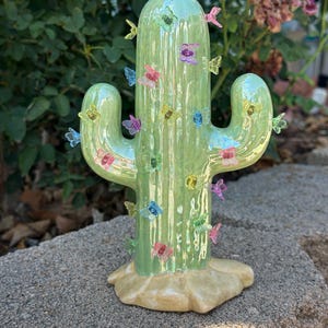 May include: A light green iridescent cactus figurine featuring a smooth, glossy surface adorned with numerous small, translucent, multi-colored butterfly-shaped accents. The cactus stands on a textured, tan-colored base shaped like a mound of earth.