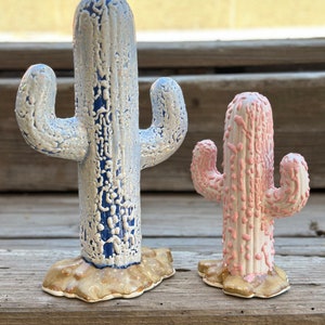 May include: Two ceramic cactus figurines, one blue and white, the other pink and white, with a textured finish. The larger cactus is taller than the smaller cactus.