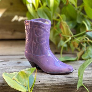May include: A purple ceramic cowboy boot planter. The boot has a pointed toe and a slight heel. The boot is sitting on a wooden surface.