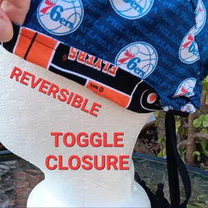 May include: A reversible face mask with a blue and orange fabric design. The blue side features a repeating pattern of the Philadelphia 76ers logo. The orange side features a black and orange basketball pattern. The mask has a toggle closure and a black drawstring.