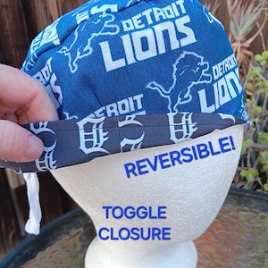 May include: A blue and black reversible baseball cap with a Detroit Lions pattern on one side and a Detroit Tigers pattern on the other side. The cap has a toggle closure and a white drawstring.