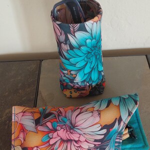 May include: Two floral print eyeglass cases. One is a tall, cylindrical case, and the other is a flat, rectangular case. Both cases feature a vibrant floral pattern with teal, pink, and orange hues on a dark background. The tall case holds a pair of glasses.