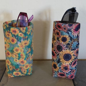 May include: Two fabric eyeglass cases with sunflower patterns. One case has a teal background with yellow and orange sunflowers. The other case has a dark blue background with colorful sunflowers. Each case holds a pair of eyeglasses.