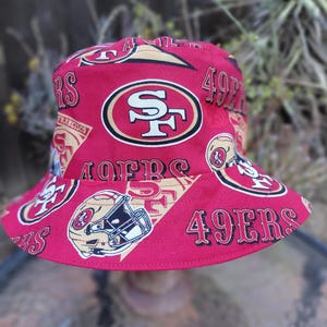 May include: Red bucket hat featuring the San Francisco 49ers logo and team name. The hat has a repeating pattern of the team's logo, helmet, and the words "49ers" in black and gold. The hat is made of a soft fabric.