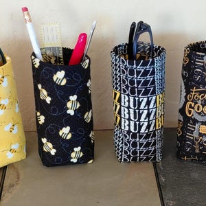 May include: Six fabric pen holders in various patterns. Designs include yellow with bees, black with bees, black and white "BUZZ" text, black with "Focus on the Good" text, and black with yellow polka dots. Some holders contain pens, pencils, and glasses.