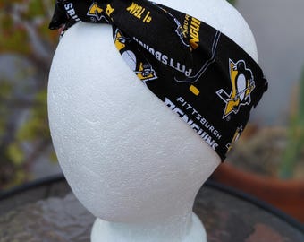 May include: Black and yellow Pittsburgh Penguins headband. The headband features the team's logo, the words "Penguins" and "Pittsburgh" in white, and hockey-related graphics. The headband is tied at the top.