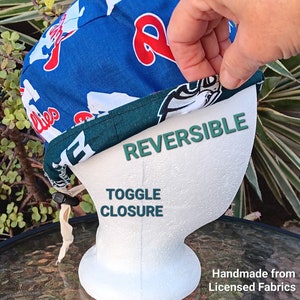 May include: Reversible baseball cap with a toggle closure. One side features a blue and white Philadelphia Phillies logo, the other side features a green and white Philadelphia Eagles logo. Handmade from licensed fabrics.