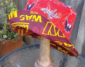 May include: A reversible bucket hat featuring a red exterior with the text "capitals" and a logo, and a maroon interior with yellow text. The hat is displayed on a wooden hat stand.