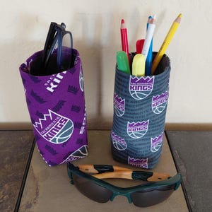 May include: Two fabric pen holders with a Sacramento Kings basketball team logo. One is purple, the other is gray. The purple holder contains black glasses. The gray holder contains pens and pencils. Green sunglasses rest in front.