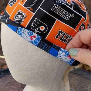 May include: A scrub cap featuring a repeating pattern of black and orange squares with Philadelphia Flyers logos, hockey sticks, and NHL emblems. The bottom edge is trimmed with a blue and white fabric band displaying Philadelphia 76ers logos.