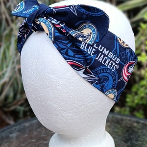 May include: A blue and white headband with a repeated pattern of the Columbus Blue Jackets logo. The logo features a hockey stick, a puck, and the words "Columbus Blue Jackets".