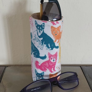 May include: A cylindrical glasses case with a white background, featuring colorful chihuahua illustrations. The case holds a pair of sunglasses and a pair of purple-framed eyeglasses. The chihuahua illustrations are in shades of blue, orange, and pink.